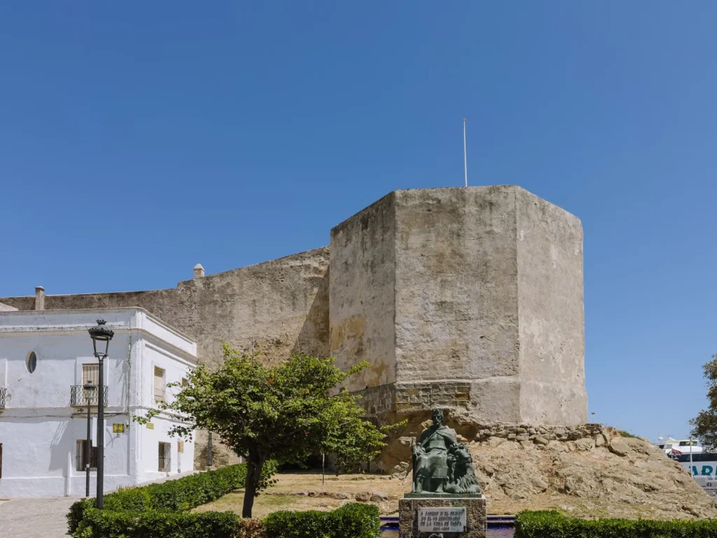 The medieval stone walls and tower of Castillo de Guzmán el Bueno in Tarifa, with a bronze statue of Guzmán el Bueno on a plinth in the foreground and a white building to the left