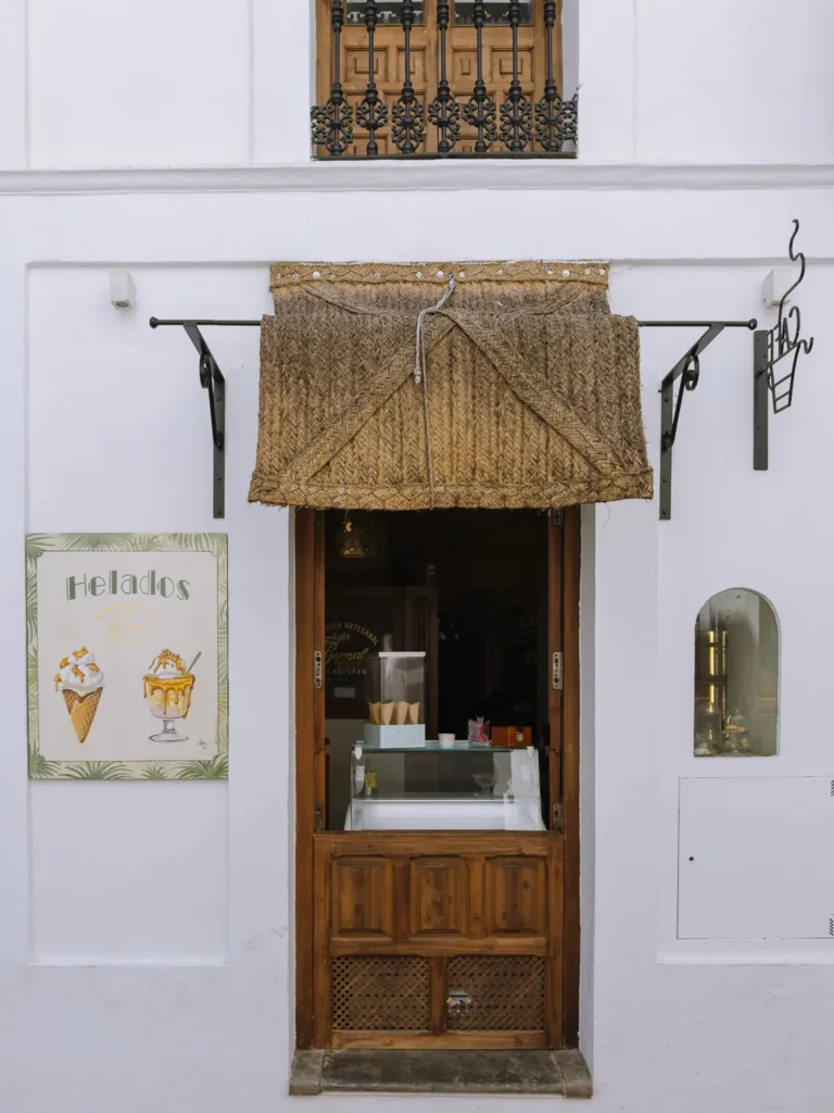 The charming entrance of an artisan ice cream shop in Tarifa, with a wicker awning, a wooden half-door, an ice cream display counter, and a painted helados sign on the white wall