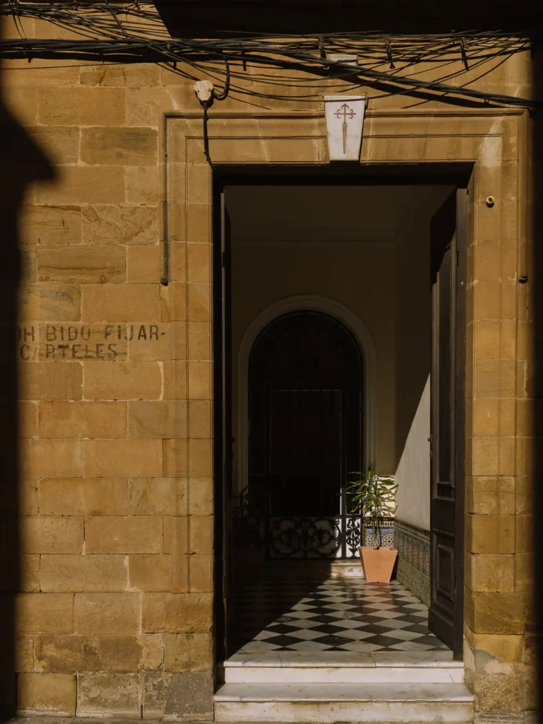 A sunlit sandstone doorway in Tarifa's old town, leading into a corridor with black-and-white checkered tiles, an ornate iron gate, and a potted plant, with a Cross of Saint James plaque above the entrance