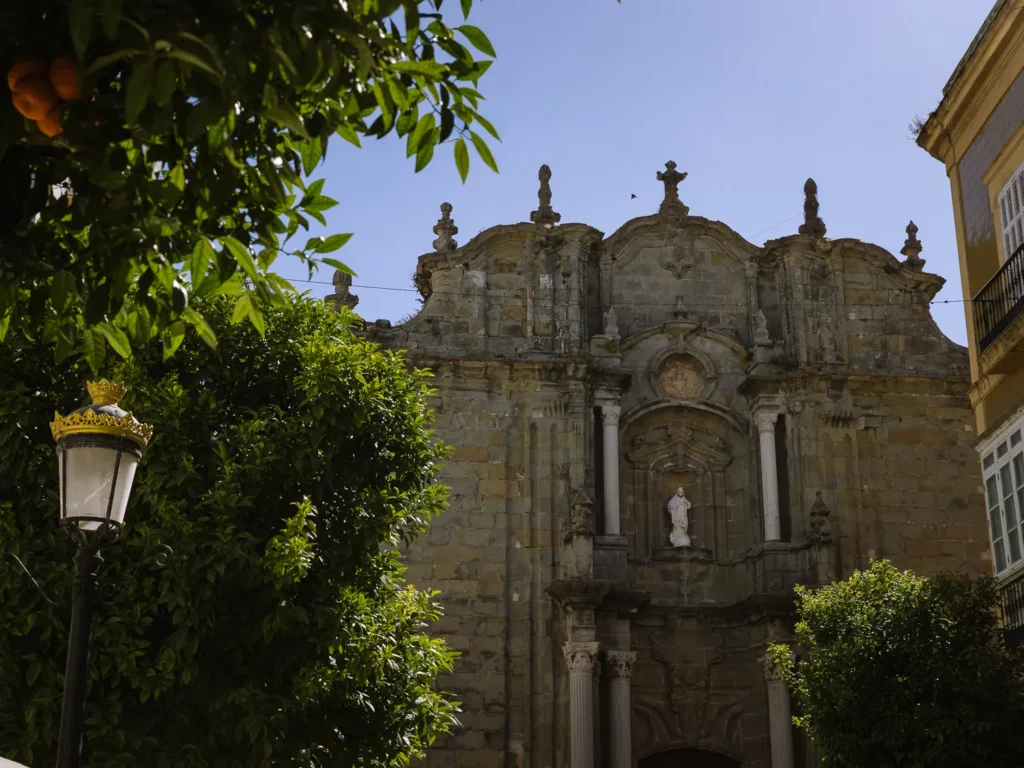 The ornate baroque stone façade of Iglesia de San Mateo in Tarifa, framed by orange trees and a decorative street lamp, with a white statue set into the entrance portal under a clear blue sky