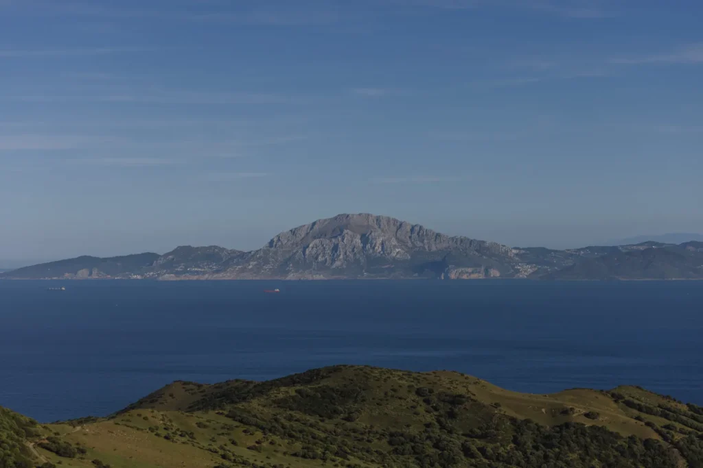 A sweeping view from the hills above Tarifa looking across the deep blue Strait of Gibraltar towards Jebel Musa in Morocco, with green rolling hills in the foreground and a cargo ship visible on the water