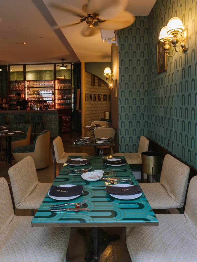 The stylish art deco interior of Palcíneo restaurant in Tarifa, with a patterned teal table set for four, cream upholstered chairs, retro arched wallpaper, brass wall sconces, and a glowing bar in the background