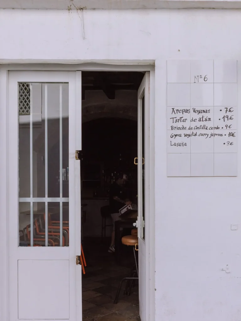 The white-walled entrance of a restaurant in Tarifa, numbered No. 6, with a handwritten tile menu outside listing dishes including tuna tartare and vegan arepas