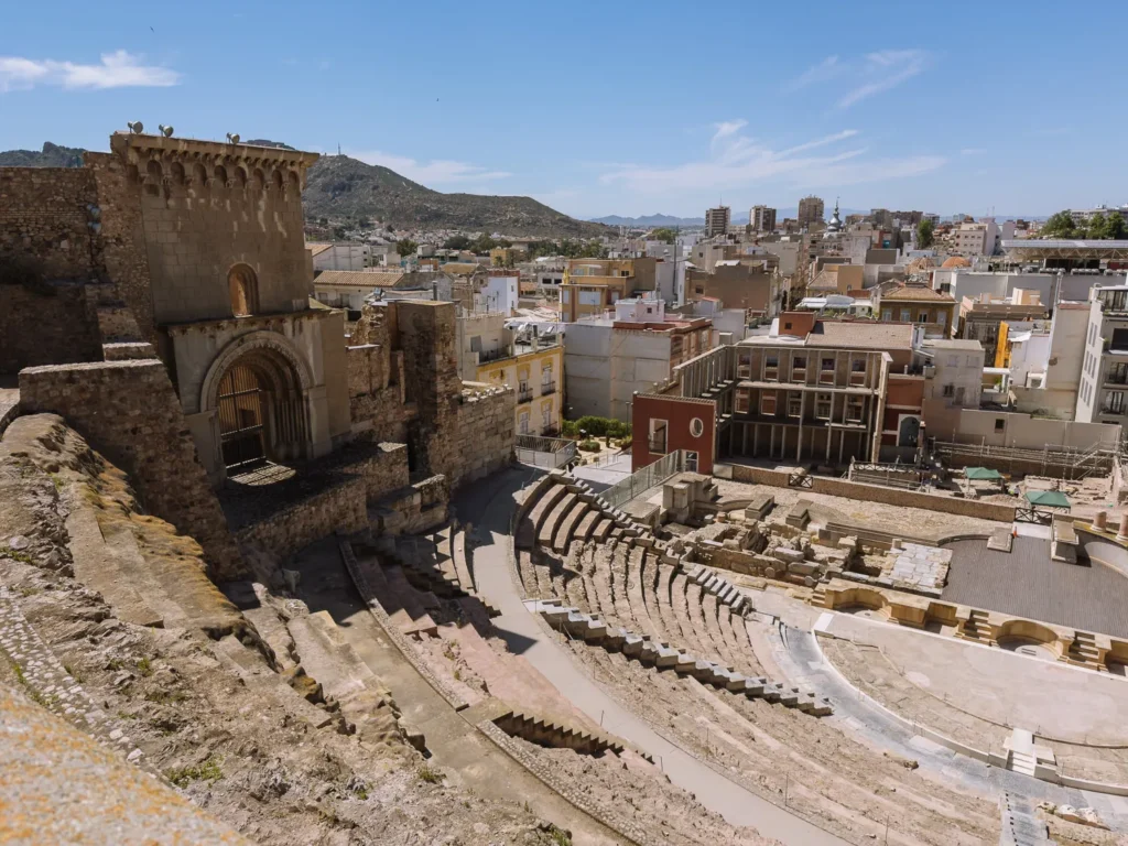 An aerial view of the Roman Theatre of Cartagena (Teatro Romano de Cartagena), a stunning 1st-century BC ruin rediscovered in 1988 and beautifully restored, set against the backdrop of Cartagena's cityscape and surrounding hills in Murcia, Spain.