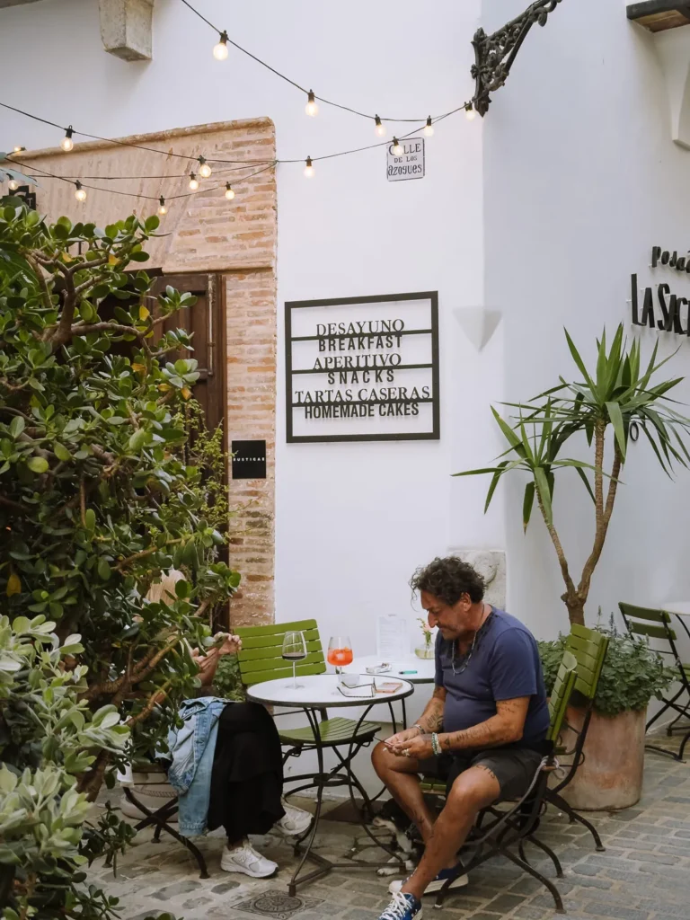 Two people relaxing with drinks at a courtyard café on Calle de los Azogues in Tarifa, surrounded by lush greenery, string lights, and a menu board listing breakfast, snacks, and homemade cakes, with the Posada La Sacristía sign visible to the right