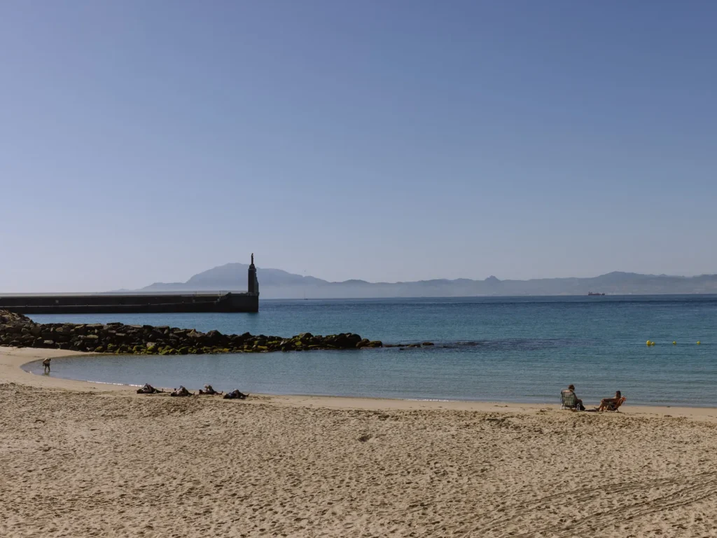 A quiet sandy beach in Tarifa with a few sunbathers and a harbour wall with a small monument, looking across the calm blue waters of the Strait of Gibraltar towards the mountains of Morocco