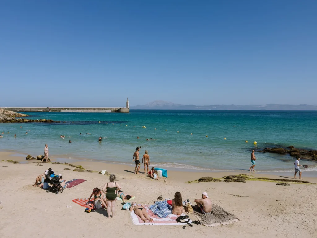 Sunbathers and swimmers on a sandy beach in Tarifa on a clear summer day, with turquoise water, a stone harbour wall, and the mountains of Morocco visible across the Strait of Gibraltar