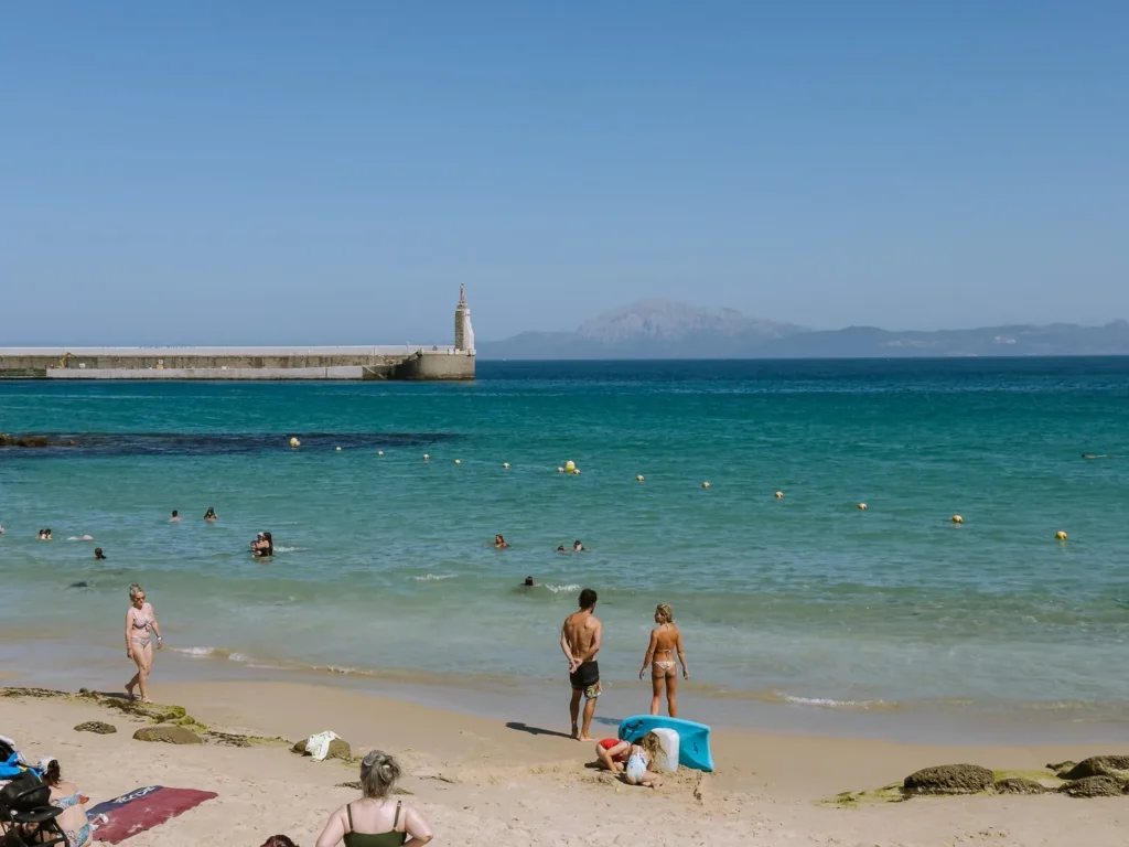 Swimmers and sunbathers on Playa Chica in Tarifa on a sunny day, with turquoise water, a long harbour wall with a small monument, and the hazy mountains of Morocco visible across the Strait of Gibraltar