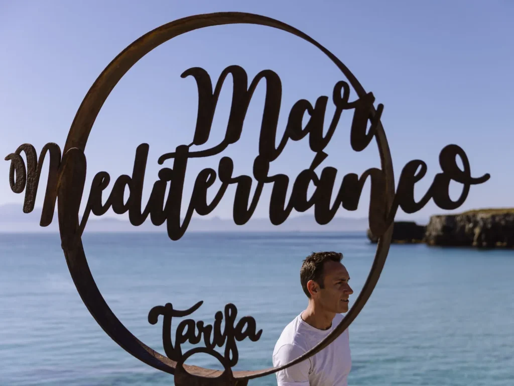 A man in a white t-shirt standing behind the circular rusty metal "Mar Mediterráneo Tarifa" sign at Tarifa Point, with turquoise sea, rocky coastline, and the hazy mountains of Morocco visible in the background