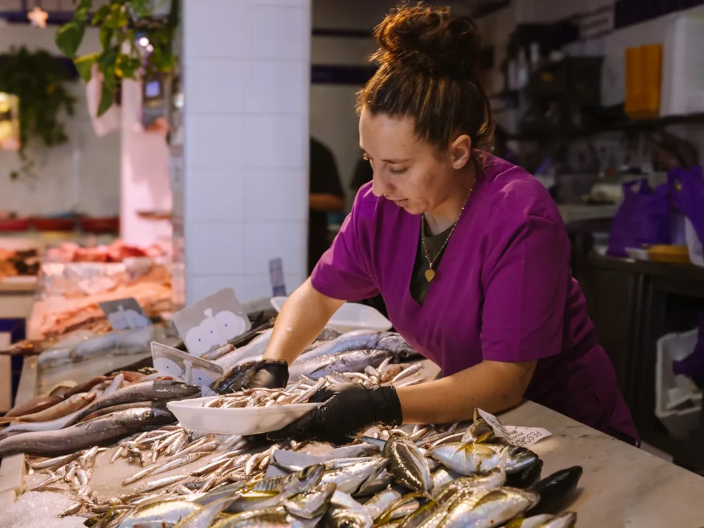 A female fish vendor in a purple apron arranging fresh fish on an ice-covered counter at Tarifa's Mercado Municipal, with a variety of whole fish and anchovies displayed in the foreground