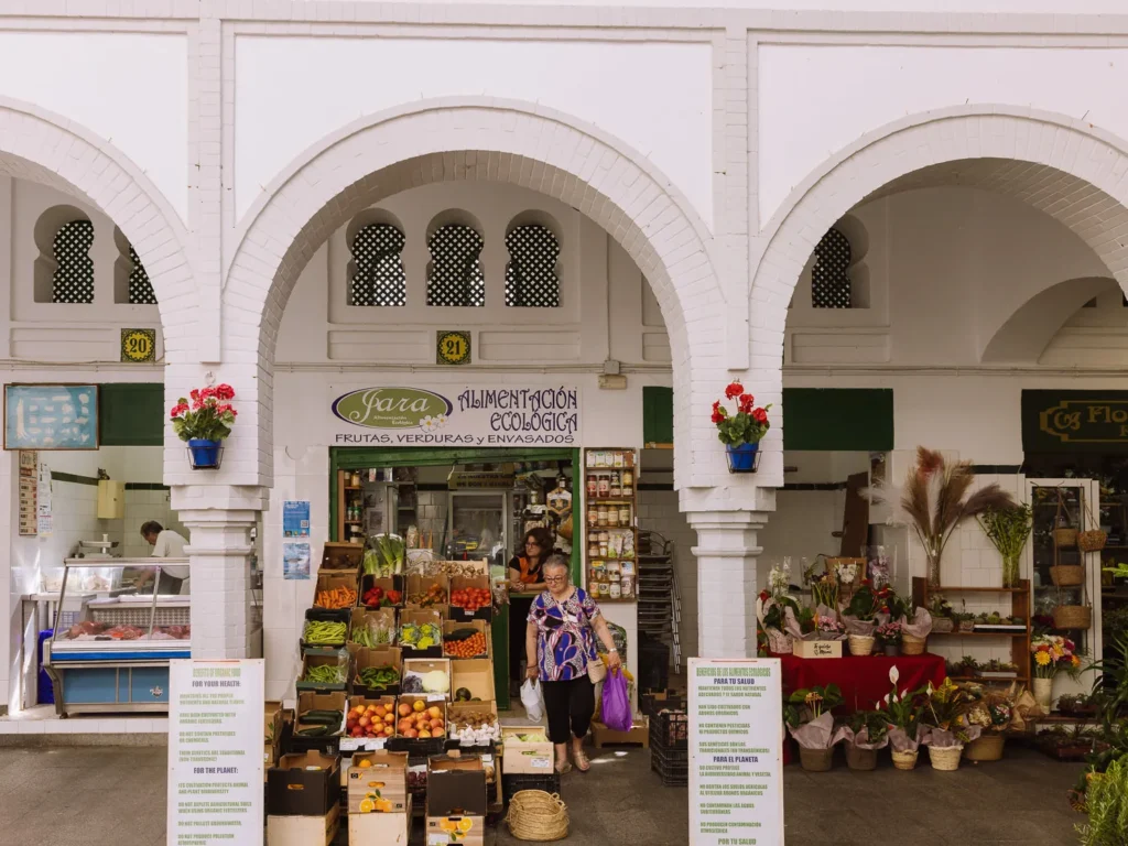 The white Moorish-arched interior of Tarifa's Mercado Municipal, with a woman leaving a stall selling organic fruit and vegetables, a flower shop to the right, and a butcher to the left