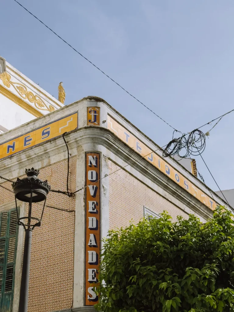 The ornate corner façade of the historic Novedades Tejidos building in Tarifa, covered in patterned terracotta tiles with blue and gold signage, a decorative roofline with a small statue, and a street lamp in the foregroun