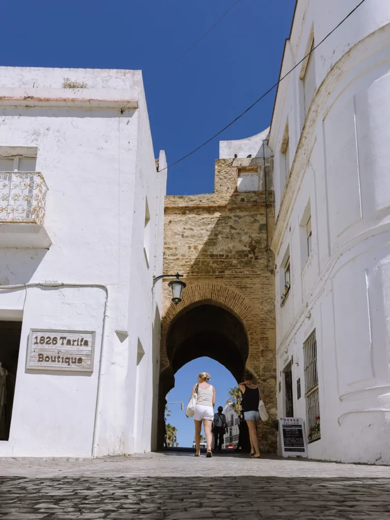Two visitors walking through a Moorish brick arch gateway into Tarifa's white-washed old town on a sunny day, with cobblestone streets and a 1826 Tarifa Boutique sign visible on the left