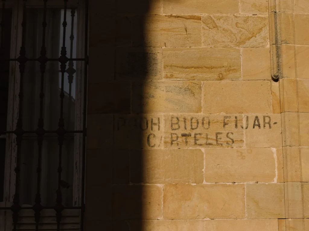 A sunlit sandstone wall in Tarifa's old town with a faded stencilled inscription reading "Prohibido Fijar Carteles" (No Bill Posting), partially in shadow beside a barred iron window