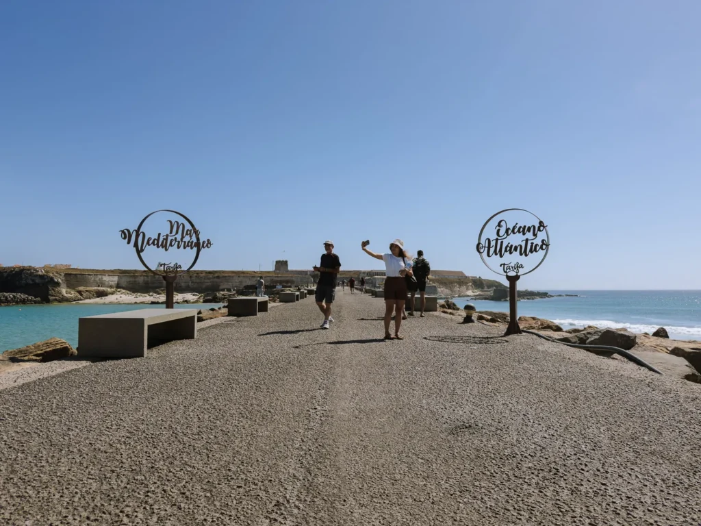Two visitors taking a selfie on the walkway at Tarifa Point, flanked by decorative metal signs marking the Mar Mediterráneo and Océano Atlántico, with turquoise water visible on both sides under a clear blue sky