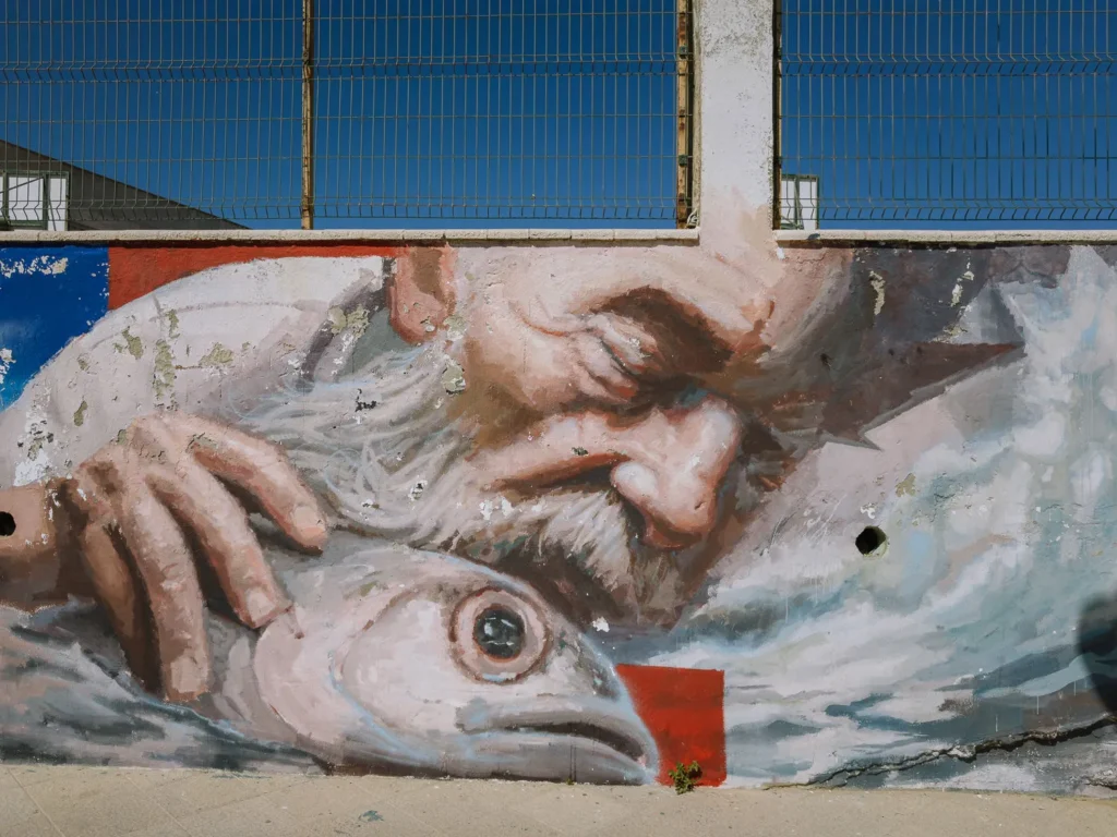 A large-scale street art mural on a wall in Tarifa depicting a weathered fisherman clutching a large tuna fish close to his face, painted in muted tones of grey, white and pink against a blue sky