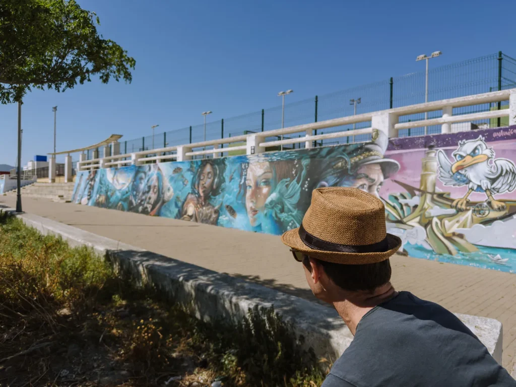 A man in a straw hat sitting on a low wall admiring a large colourful street art mural on Tarifa's seafront, depicting underwater figures, mermaids and a seagull character under a clear blue sky