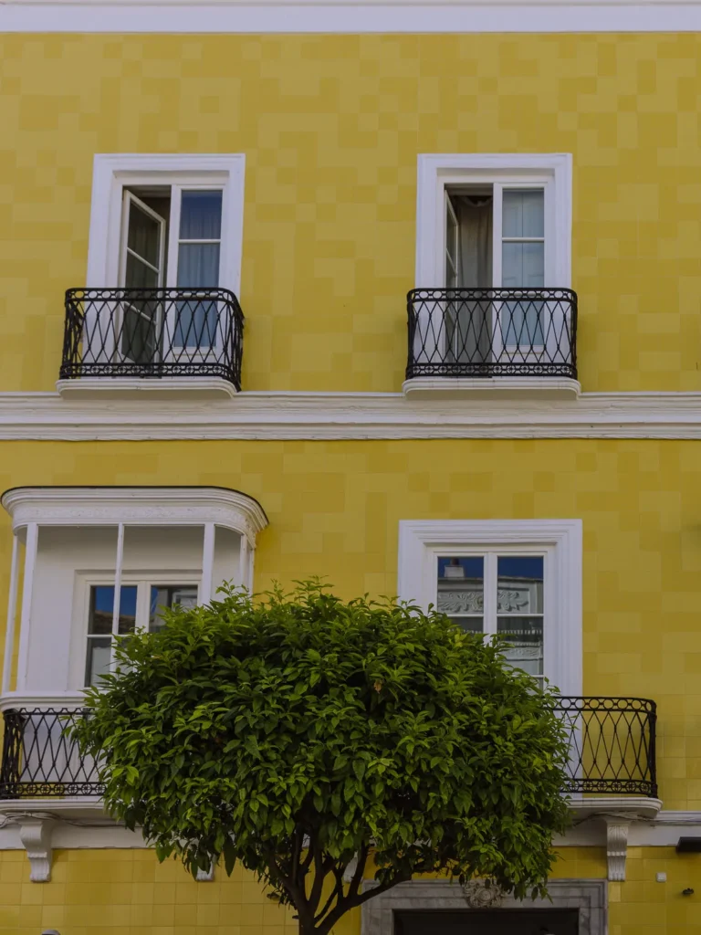 The vibrant yellow tiled façade of a building in Tarifa with white-framed windows, ornate black wrought iron balconies, and a neatly pruned green tree in the foreground