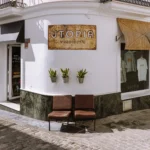 The exterior of Utopia, a boutique clothing store in Tarifa's white-washed old town, with a rustic metal sign, wicker awnings, potted snake plants, and two leather chairs on a cobblestone street