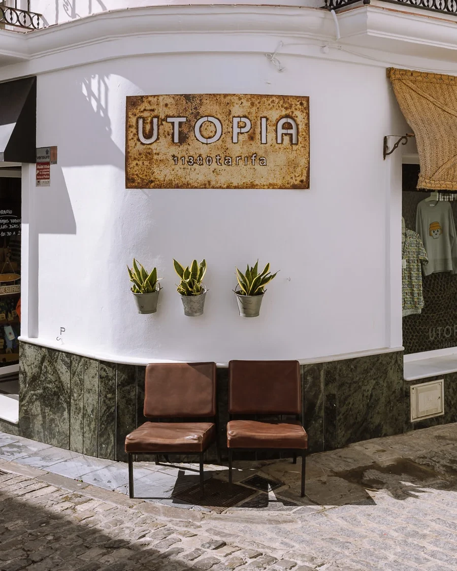 The exterior of Utopia, a boutique clothing store in Tarifa's white-washed old town, with a rustic metal sign, wicker awnings, potted snake plants, and two leather chairs on a cobblestone street