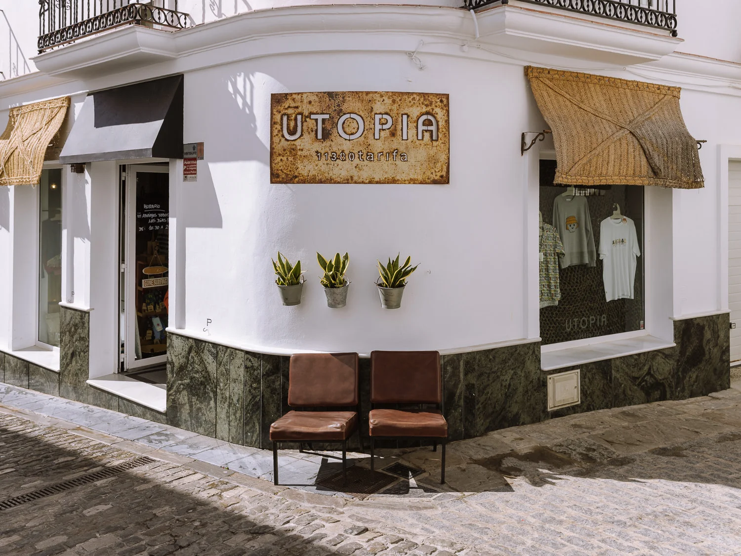 The exterior of Utopia, a boutique clothing store in Tarifa's white-washed old town, with a rustic metal sign, wicker awnings, potted snake plants, and two leather chairs on a cobblestone street