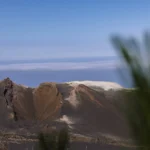 Volcanic crater landscape on La Palma in the Canary Islands, with rugged dark slopes, a pale ridge, and ocean clouds in the distance.