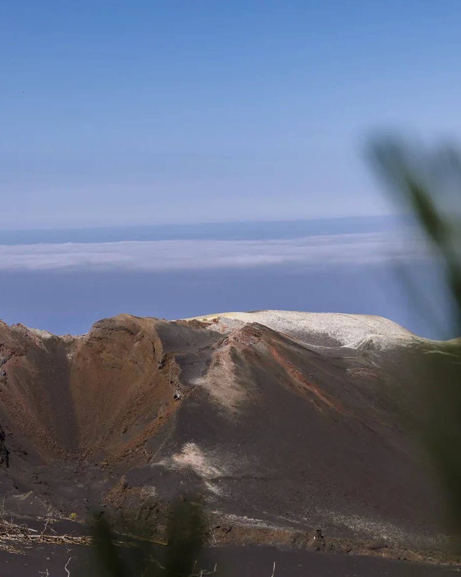 Volcanic crater landscape on La Palma in the Canary Islands, with rugged dark slopes, a pale ridge, and ocean clouds in the distance.