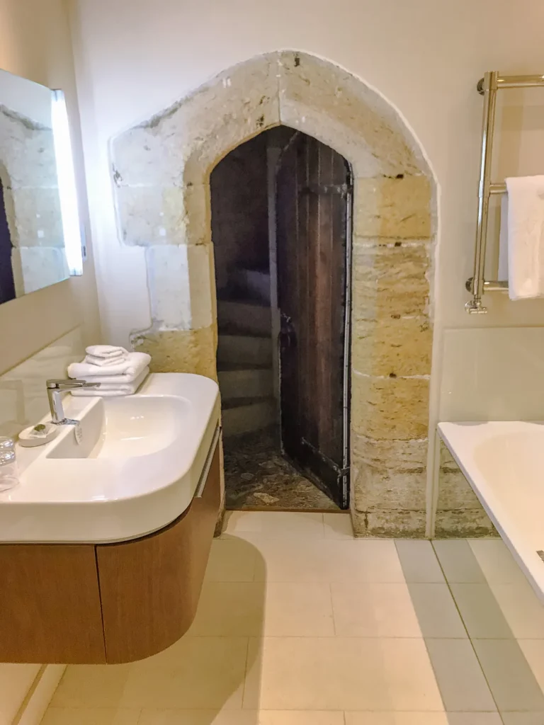 Modern bathroom sink beside an original stone archway with a wooden door leading to a turret staircase at Amberley Castle