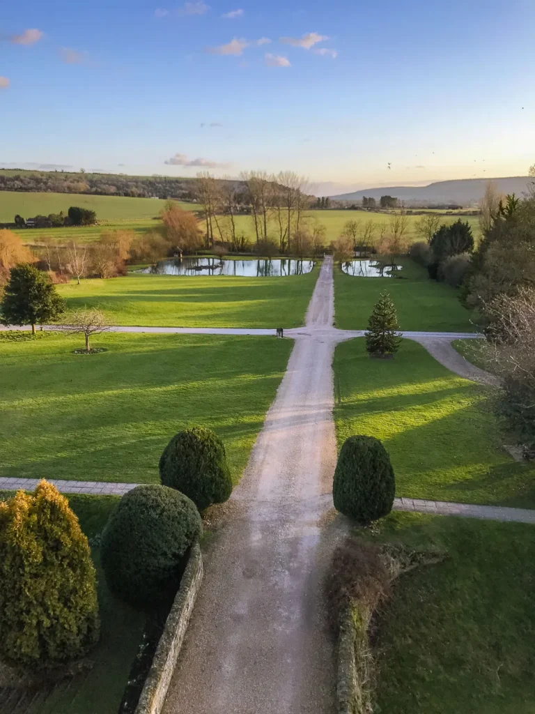 Gravel driveway leading through landscaped grounds to a pond, with green fields and the South Downs beyond near Amberley, England.