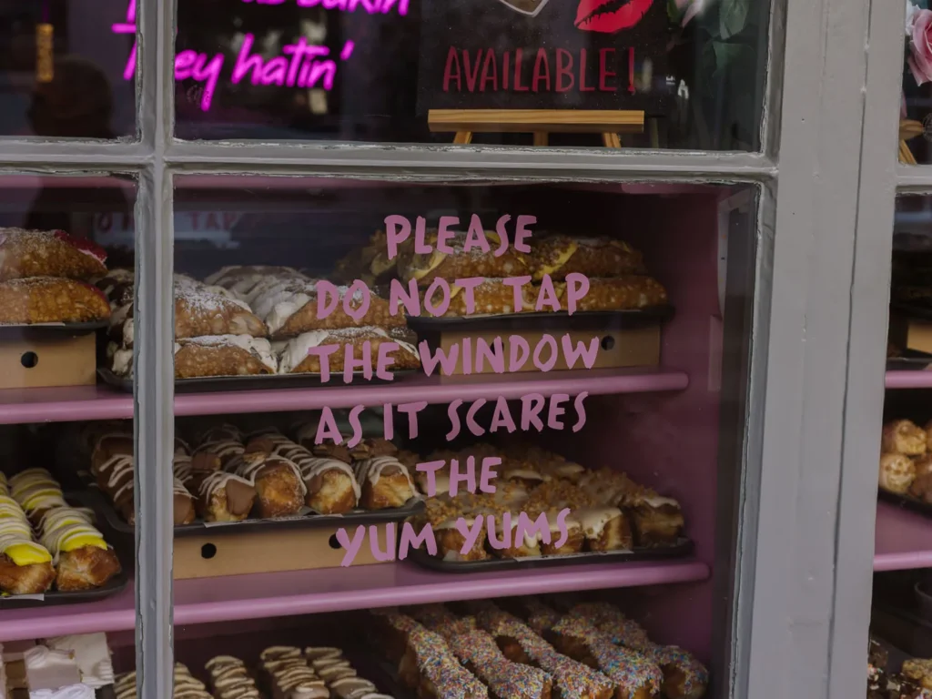Trays of iced and sugared doughnuts fill a bakery window, with pink lettering reading “Please do not tap the window as it scares the yum yum”.