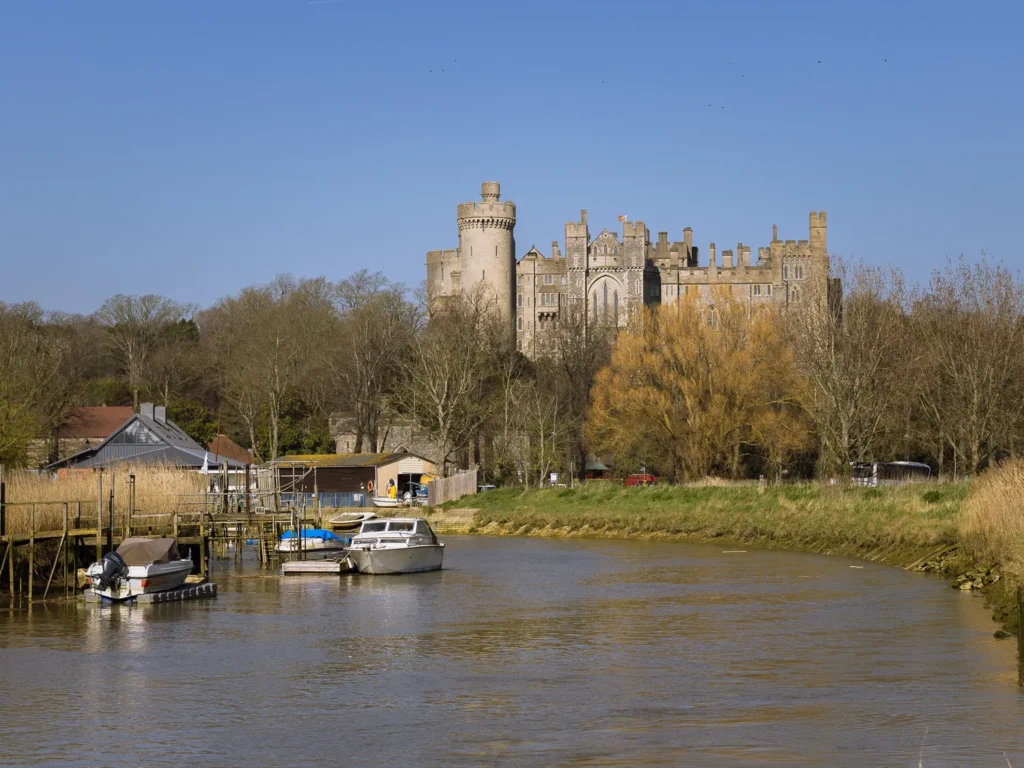 View along the River Arun towards Arundel Castle, with small motorboats moored beside a wooden jetty and leafless riverside trees framing the castle’s towers.