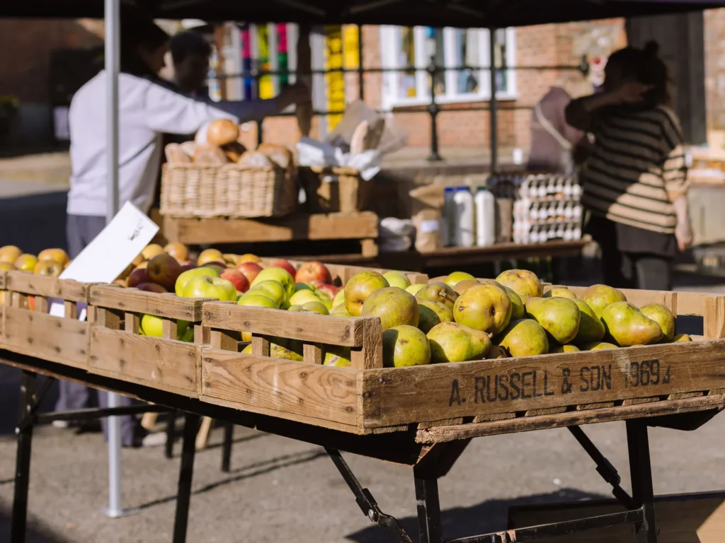 Wooden crates filled with green pears and red apples on a market stall, with bread baskets, bottled drinks and shoppers in the background at Arundel Farmers Market.