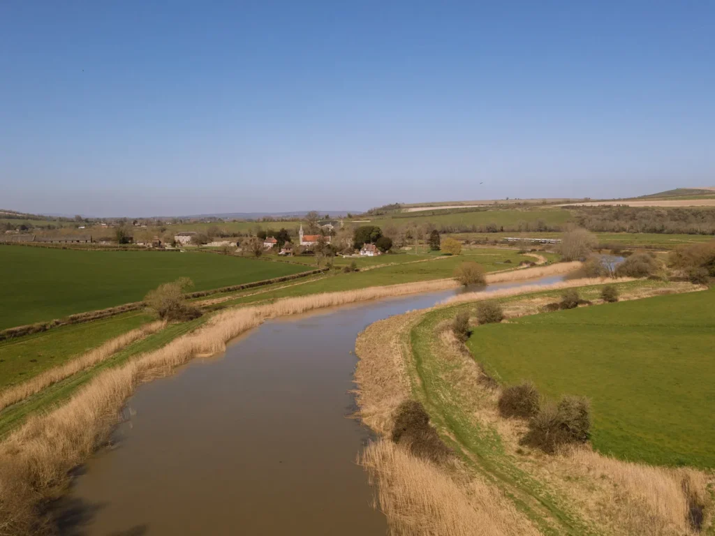 Aerial view of the River Arun winding through green fields and reed beds near Amberley, with the village and rolling South Downs countryside in the distance.