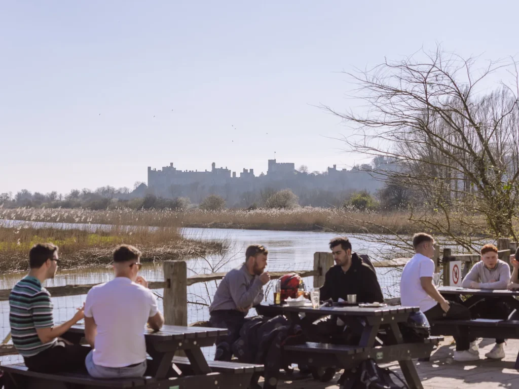 People at picnic tables in a riverside beer garden, with a hazy castle skyline visible across the water