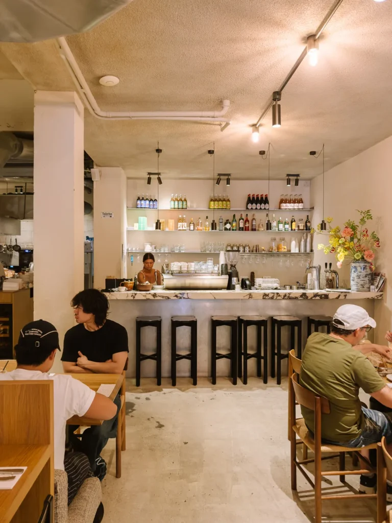 Customers seated inside a softly lit Amsterdam café with a bar counter, shelves of bottles and a staff member behind the counter