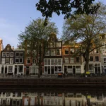 Tall canal houses with large trees along a quiet Amsterdam canal, reflected in the water at sunset