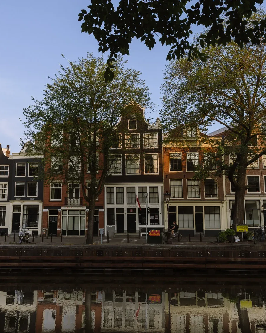 Tall canal houses with large trees along a quiet Amsterdam canal, reflected in the water at sunset
