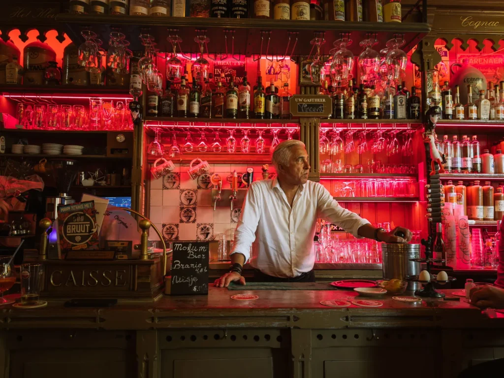 Bartender in a white shirt stands behind a dark wooden bar in an Amsterdam brown café, surrounded by red-lit shelves of bottles, hanging glasses and beer taps.
