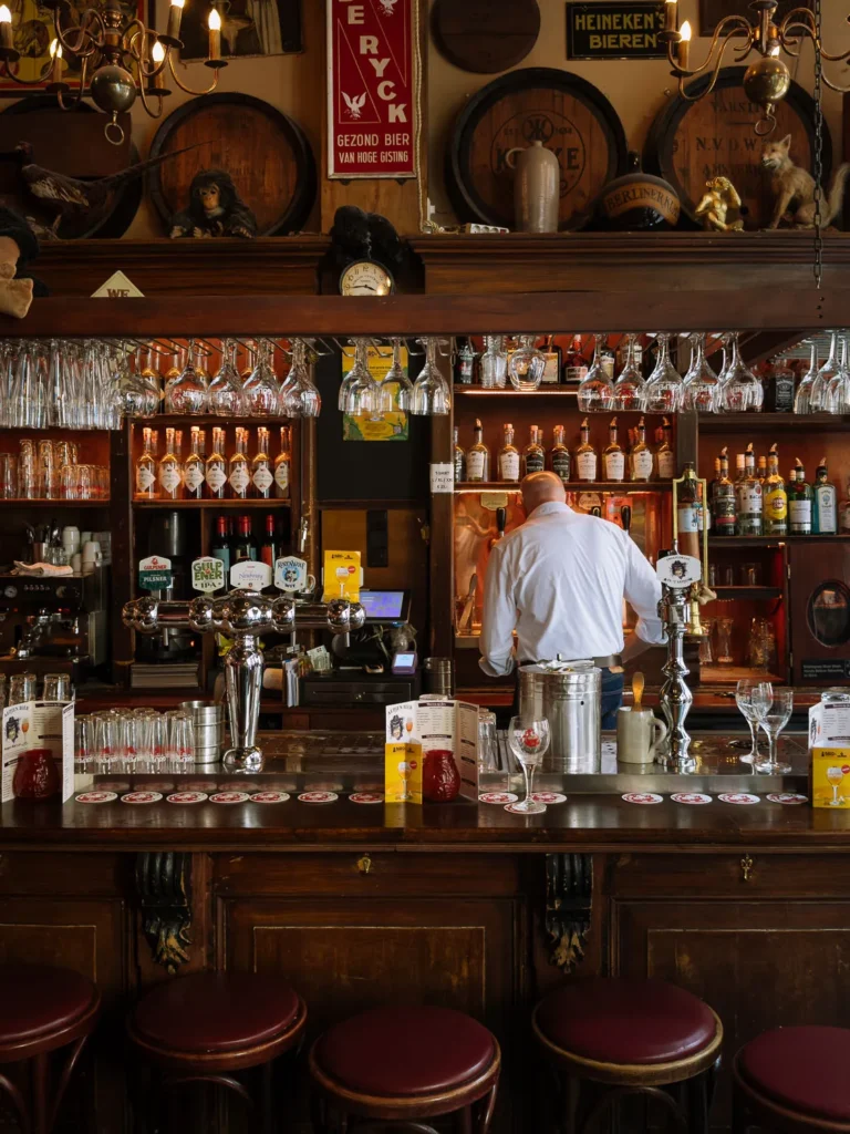 Bartender seen from behind works at a polished wooden bar in an Amsterdam brown café, with rows of bottles, hanging wine glasses and old beer barrels decorating the wall above.