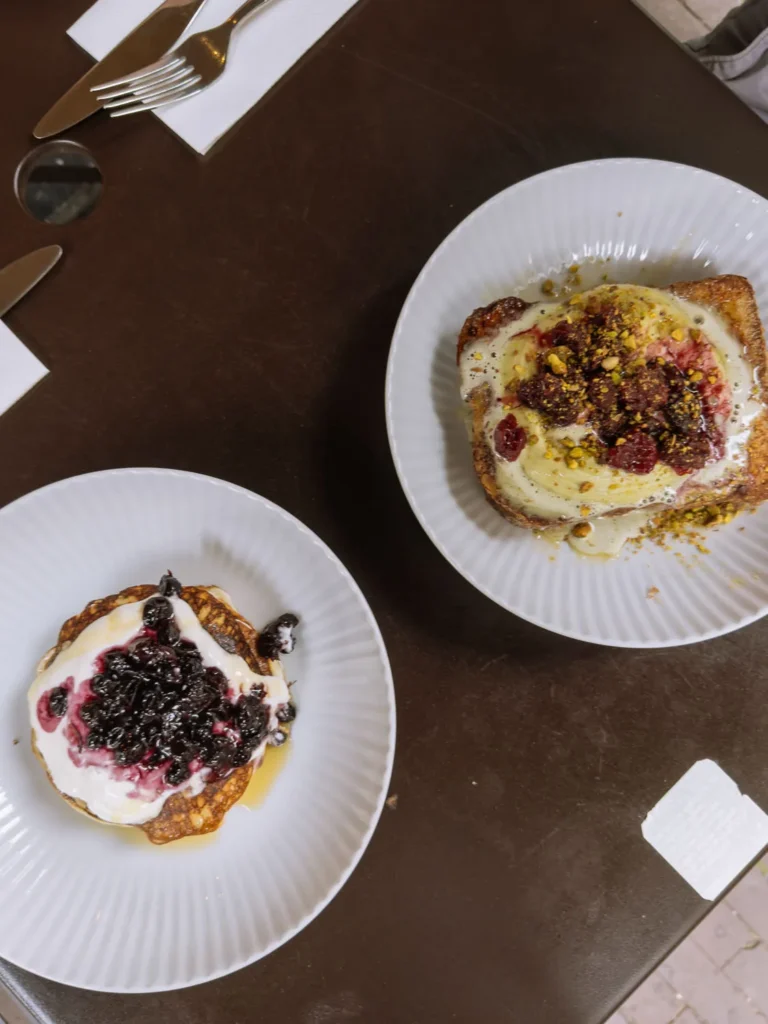 Two plates of toast topped with ricotta, berries and pistachios on a dark café table in Amsterdam