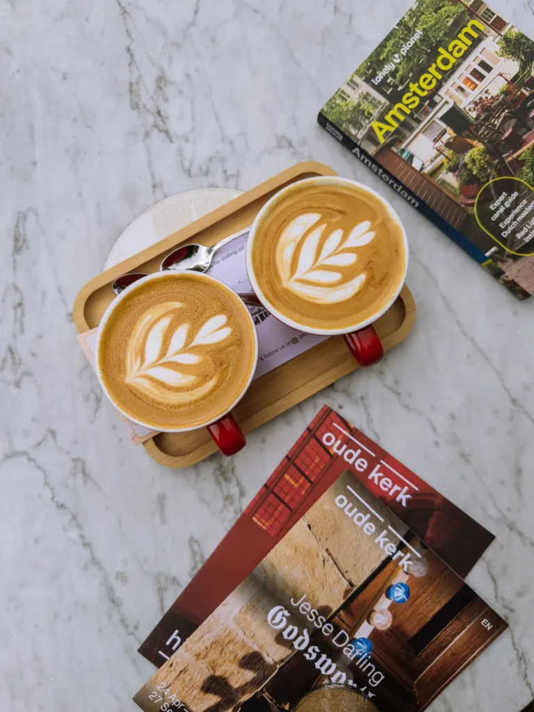 Two red cups of latte with leaf-shaped foam art on a wooden tray beside Amsterdam guidebooks on a white marble table