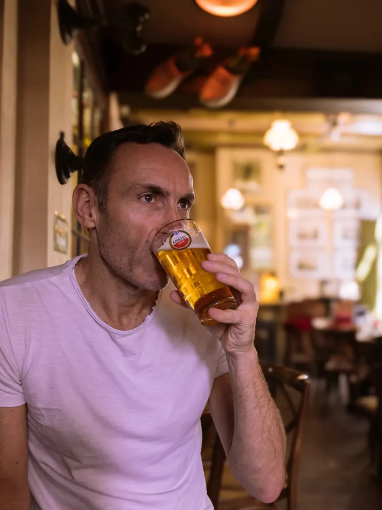 Man in a light T‑shirt drinks a glass of golden beer inside a cosy Amsterdam brown café, with wooden chairs, soft lamps and framed pictures blurred in the background.