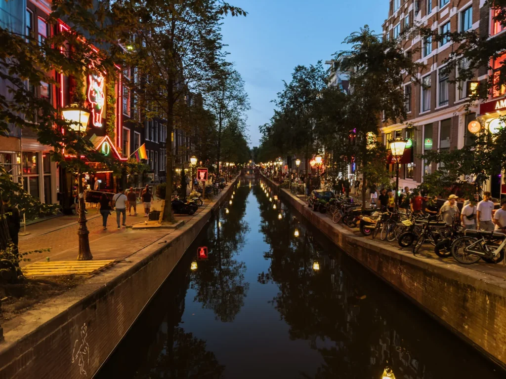 Evening scene along a narrow canal in Amsterdam’s Red Light District, with neon signs, rainbow flags, trees and crowds of people reflected in the dark water between brick quays lined with bicycles.