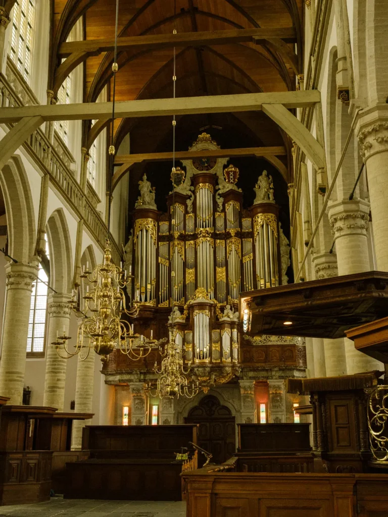 Ornate gold and dark-wood Vater-Müller organ rising behind brass chandeliers, pale stone columns and the timber vault of Amsterdam’s Oude Kerk