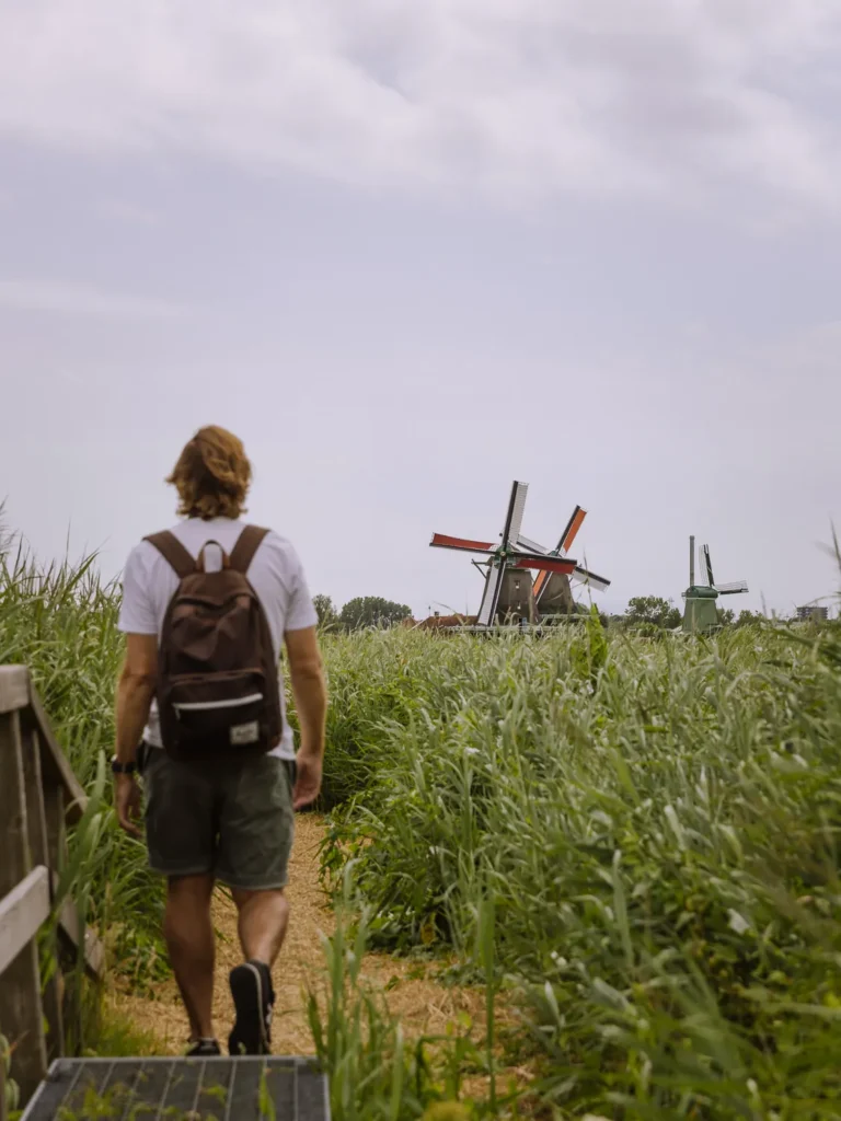 Person with a backpack walks along a grassy path toward windmills at Zaanse Schans under a cloudy sky