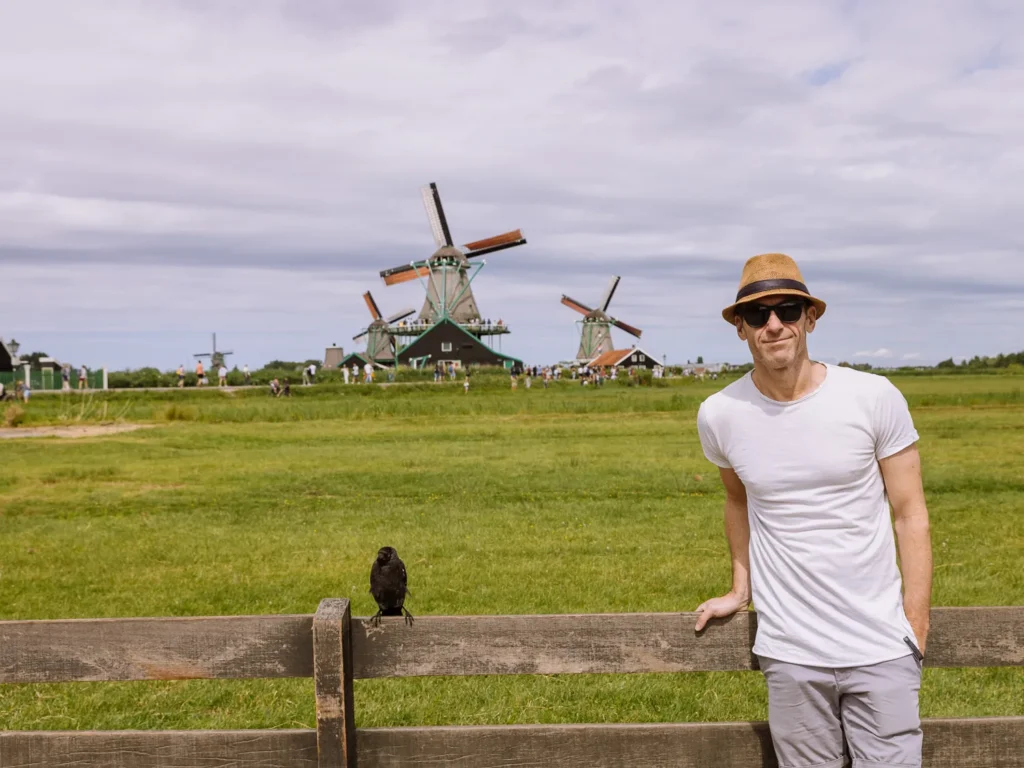 Man in a straw hat poses in front of windmills on the Zaanse Schans with a bird perched on the fence beside him
